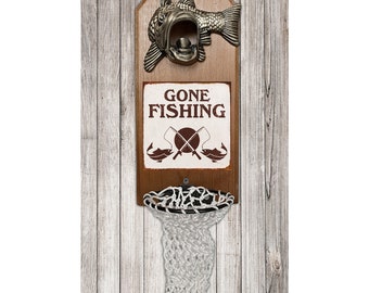 Op de afbeelding: Een houten, aan de muur gemonteerde flessenopener met een visontwerp. De opener heeft een net om flessenkurken op te vangen. Het bord zegt "Gone Fishing" met een hengel en twee vissen.