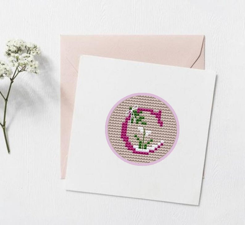 May include: A white card with a pink envelope features a cross-stitch design. The design is a letter "G" in a circle, with a floral motif in pink, green, and white. The background is a light tan color.