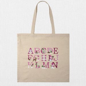 May include: A beige canvas tote bag featuring a cross-stitch alphabet design in shades of pink and white. The letters A through M are displayed with decorative elements. The bag has two handles for carrying.