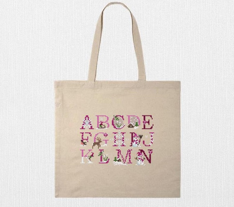 May include: A beige canvas tote bag featuring a cross-stitch alphabet design in shades of pink and white. The letters A through M are displayed with decorative elements. The bag has two handles for carrying.
