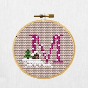 May include: A cross-stitch embroidery hoop featuring the letter "N" in a magenta color, with a small green tree and white accents. The background is a light brown fabric, and the hoop is made of wood.