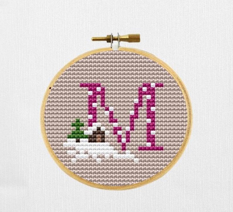 May include: A cross-stitch embroidery hoop featuring the letter "N" in a magenta color, with a small green tree and white accents. The background is a light brown fabric, and the hoop is made of wood.