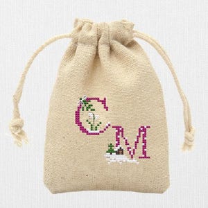 May include: A beige burlap drawstring bag with a cross-stitched design. The design features the letters "CM" in a magenta color, with floral accents. The bag has a natural, rustic appearance.