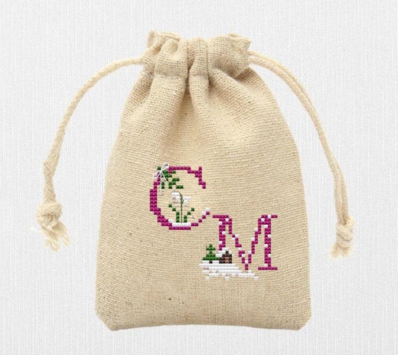 May include: A beige burlap drawstring bag with a cross-stitched design. The design features the letters "CM" in a magenta color, with floral accents. The bag has a natural, rustic appearance.