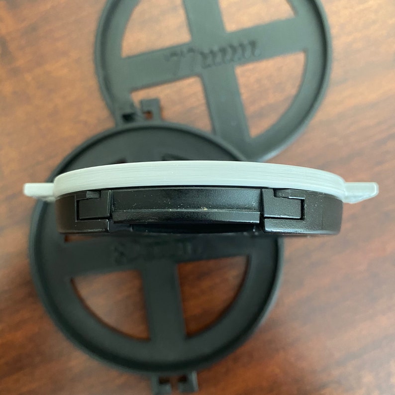 May include: A black plastic lid with a gray rim and two black clips on either side. The lid is designed to fit on a round container.