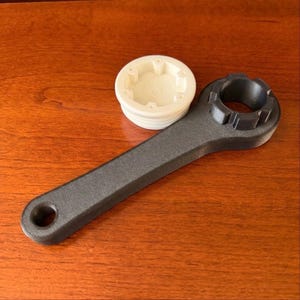 May include: A black wrench with a circular opening at one end and a round, toothed head at the other. Beside it is a white, circular cap with a series of small openings. The objects are on a wooden surface.
