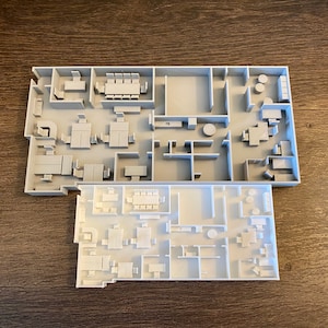 May include: Two architectural models of a building layout, one gray and one white, are displayed on a wooden surface. The models showcase rooms with furniture, including tables and chairs, offering a detailed representation of the interior design.