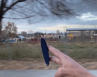 May include: A hand holding a dark blue oval-shaped object with a string attached to it. The object is being held in front of a blurry background of trees and a field.
