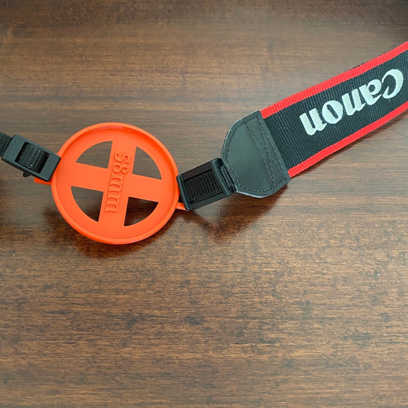May include: An orange camera lens cap with a black strap attached. The lens cap has a cross-shaped opening and the text "58mm" printed on it. The strap is labeled "Canon" in white text on a black background with a red stripe.