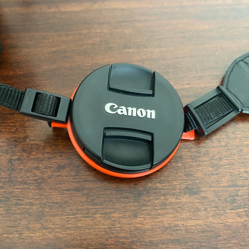 May include: A black camera lens cap with an orange rim and the word "Canon" printed in white on the top. The lens cap is attached to a black camera strap.