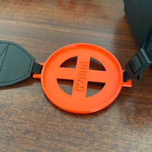May include: Orange camera lens filter cap with a 67mm marking. The cap is attached to a black camera strap.