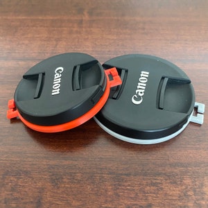 May include: Two black camera lens caps with the word "Canon" printed in white on each cap. One cap has a red and orange rim, while the other has a gray rim.
