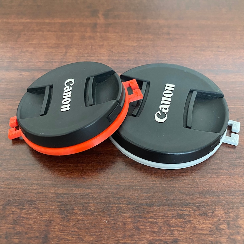 May include: Two black camera lens caps with the word "Canon" printed in white on each cap. One cap has a red and orange rim, while the other has a gray rim.