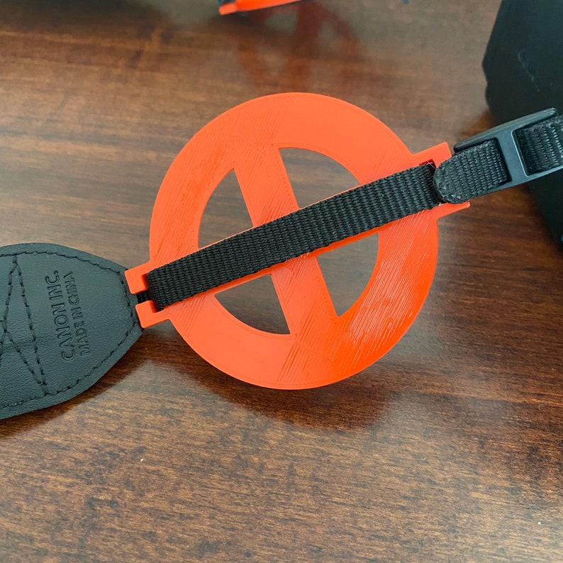 May include: A black camera strap with a red, circular, 3D printed attachment. The attachment has a cross-shaped opening in the center. The strap is labeled "Canon Inc. Made in China."