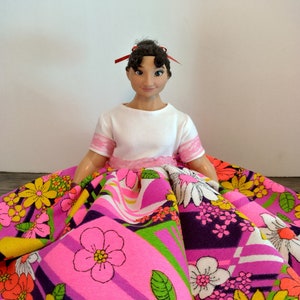 May include: A doll with dark brown hair wearing a white shirt and a pink and purple skirt with a floral pattern.