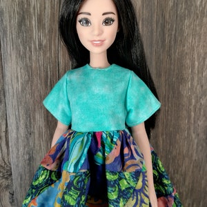 May include: A doll wearing a turquoise short-sleeved shirt and a multi-colored patchwork skirt with a floral pattern.