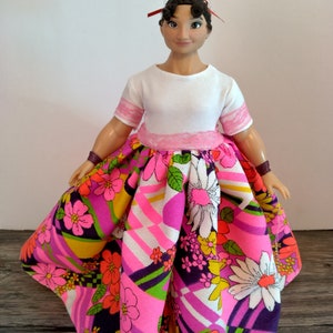 May include: A doll wearing a white shirt with pink trim and a colorful floral skirt with geometric patterns.
