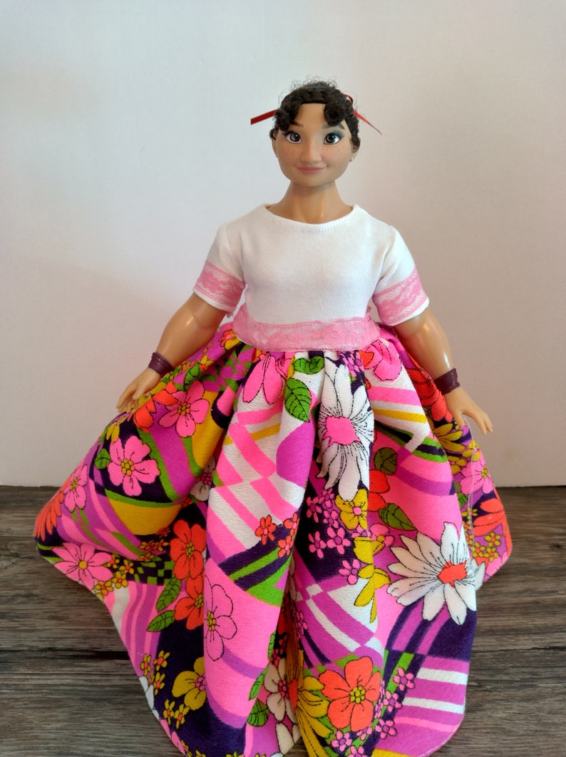 May include: A doll wearing a white shirt with pink trim and a colorful floral skirt with geometric patterns.