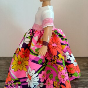 May include: A doll wearing a white top with pink trim and a colorful floral skirt. The skirt has a pink, purple, green, and white pattern with large flowers.