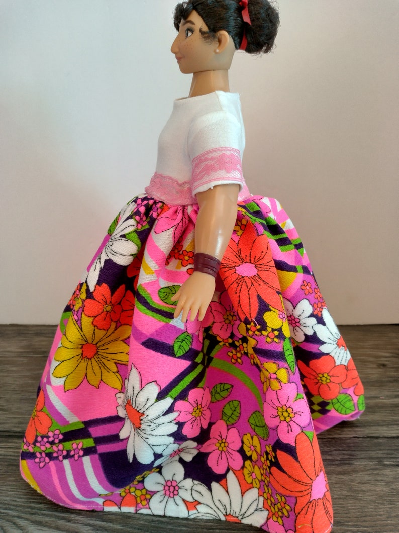 May include: A doll wearing a white top with pink trim and a colorful floral skirt. The skirt has a pink, purple, green, and white pattern with large flowers.