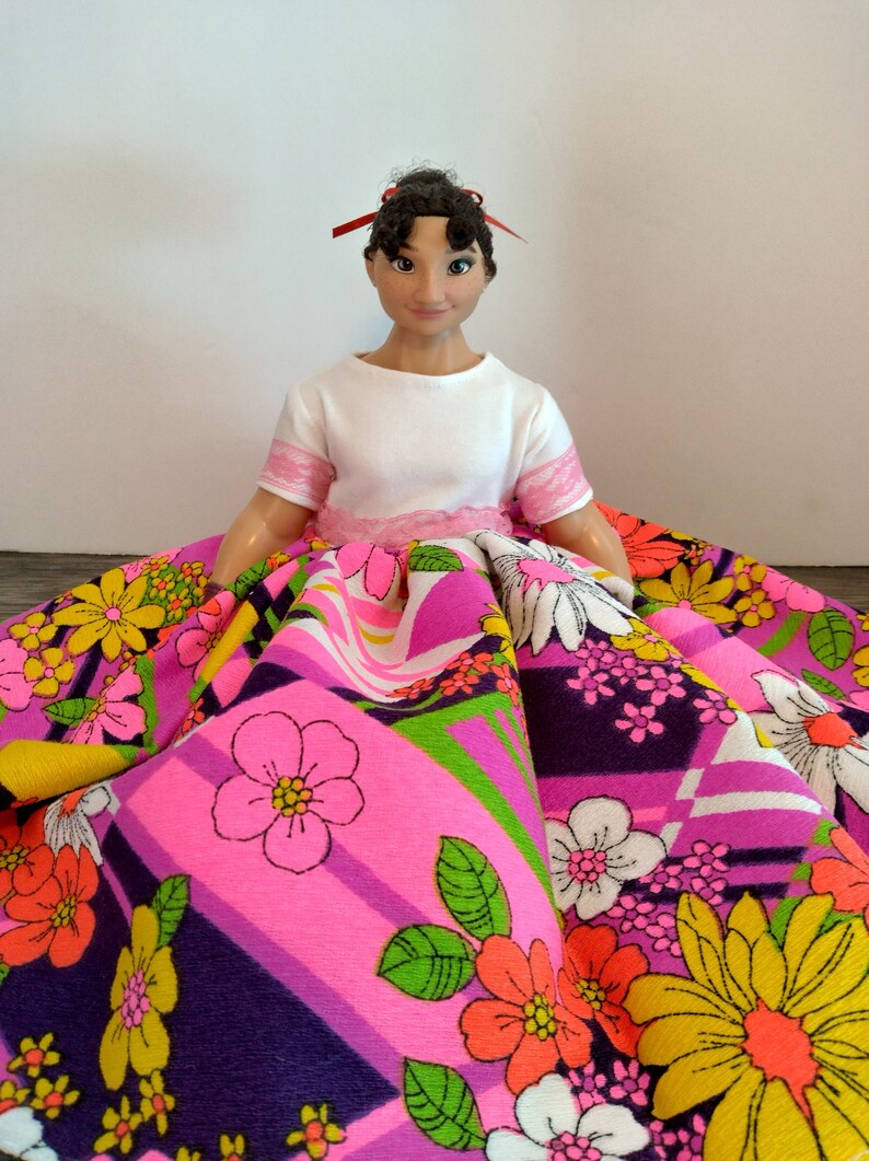 May include: A doll with dark brown hair wearing a white shirt and a pink and purple skirt with a floral pattern.