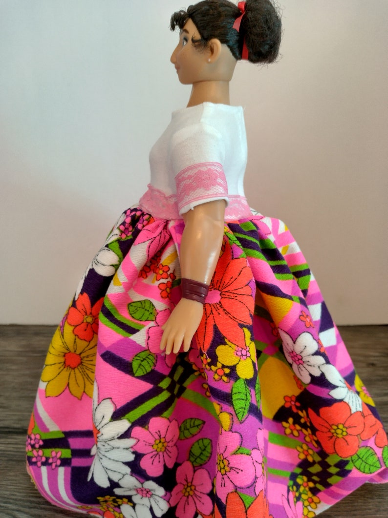 May include: A doll wearing a white blouse with pink trim and a colorful floral skirt. The skirt has a pink, green, yellow, and white pattern with large flowers.