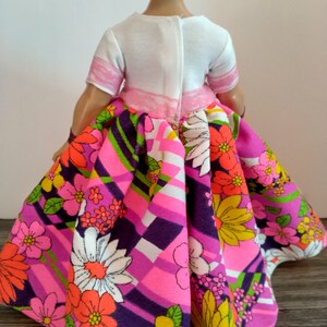 May include: A doll wearing a white top with pink trim and a pink and purple skirt with a floral pattern.