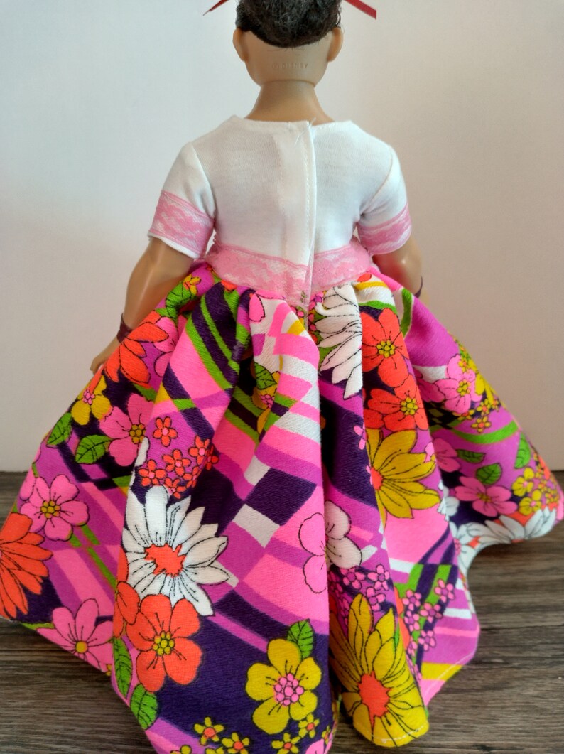 May include: A doll wearing a white top with pink trim and a pink and purple skirt with a floral pattern.