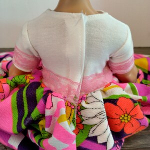May include: A doll wearing a white short-sleeved shirt with a pink lace trim and a colorful floral print skirt. The doll's neck has a "&copy; DISNEY" label.