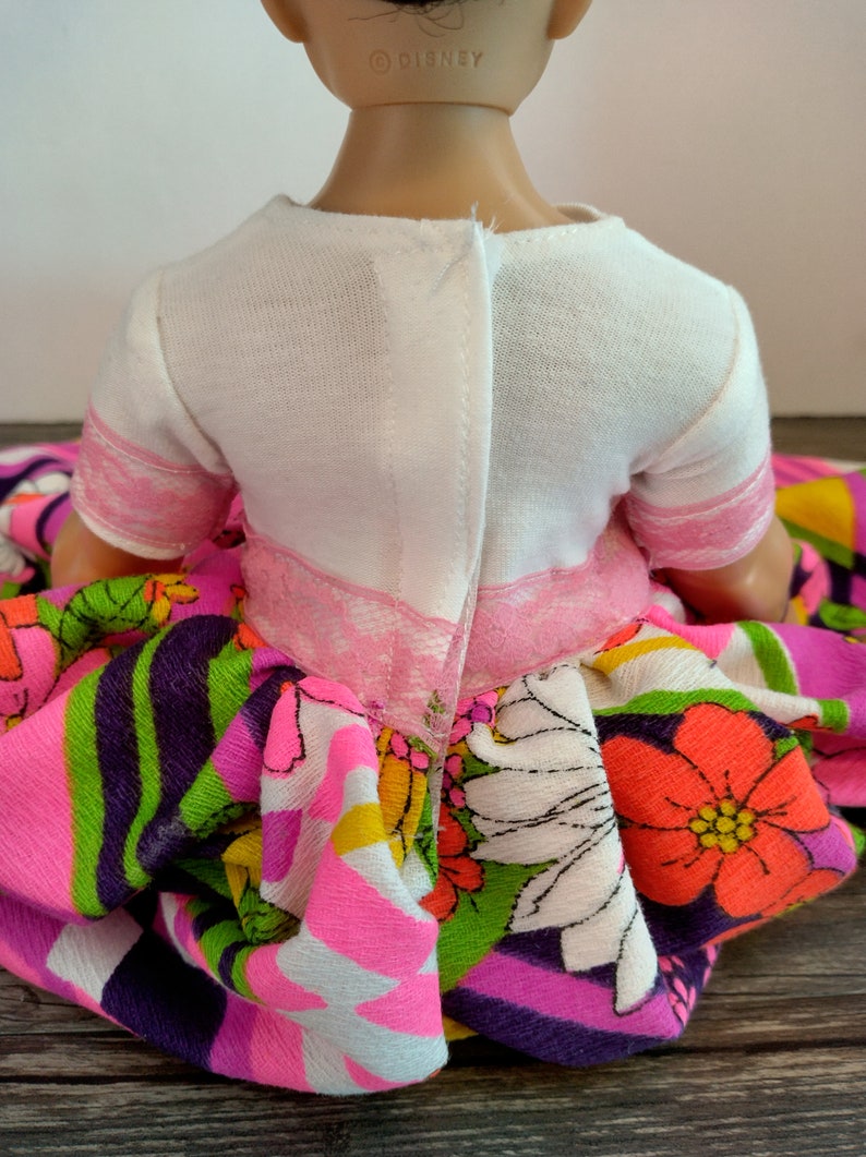 May include: A doll wearing a white short-sleeved shirt with a pink lace trim and a colorful floral print skirt. The doll's neck has a "&copy; DISNEY" label.