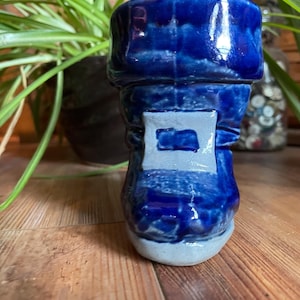 Puede incluir: Un macetero de cerámica azul en forma de bota con un detalle de glaseado blanco. El macetero tiene una superficie texturizada y una base redondeada.