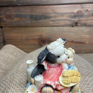 Puede incluir: Una figurita de vaca negra y blanca vestida con un vestido rosa y sosteniendo un plato de pan de maíz amarillo. La vaca está sentada en una colcha de retazos con una margarita blanca.