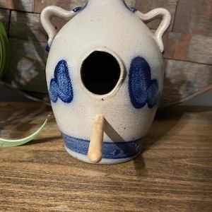 Op de afbeelding: Een blauw en wit keramisch vogelhuisje in de vorm van een kruik met twee handvatten. Het vogelhuisje heeft een rond gat waar vogels kunnen in- en uitvliegen en een houten pin om op te zitten.