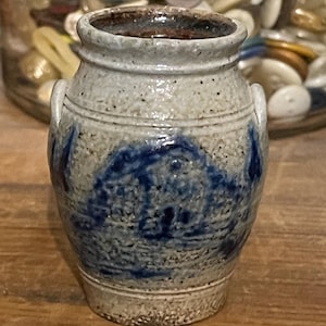 May include: A small, gray stoneware crock with blue and white painted designs. The crock has two handles and a rounded body.