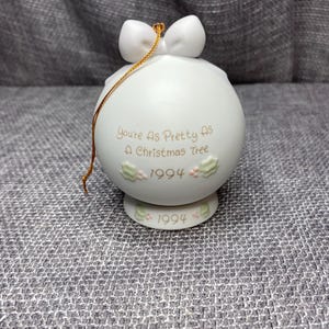 May include: White porcelain Christmas ornament with a gold ribbon and the text "You're as pretty as a Christmas tree 1994".
