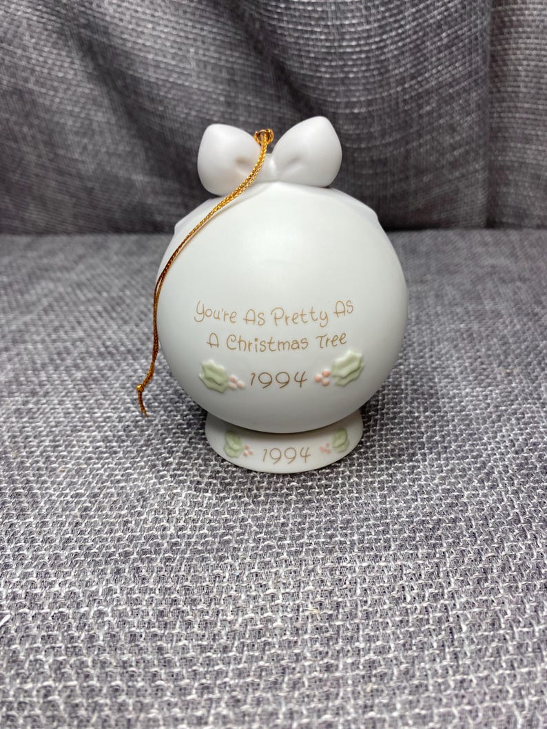 May include: White porcelain Christmas ornament with a gold ribbon and the text "You're as pretty as a Christmas tree 1994".
