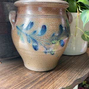 May include: A brown stoneware crock with a blue and green floral design. The crock has two handles and a textured surface.