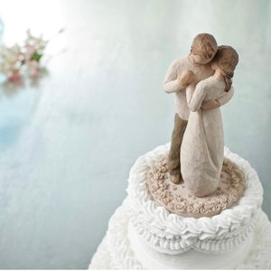 May include: A wedding cake topper featuring a couple embracing. The figures are carved from a light-colored material, with the woman in a flowing dress. The cake is white with decorative frosting and a floral base for the topper.