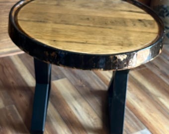 May include: A round side table featuring a light brown wooden top with a dark, weathered circular rim and two sturdy black metal legs. The tabletop surface displays natural wood grain patterns and a small white label on the edge.