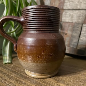 Pode incluir: Uma caneca de cerâmica marrom com alça e um design texturizado e nervurado. A caneca tem um esmalte marrom claro na metade inferior.