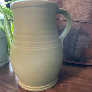 May include: A light green ceramic pitcher with a rounded body and a handle. The pitcher has a wide base and a slightly flared rim. The surface has horizontal ridges and a matte finish. The pitcher is likely handmade and suitable for serving beverages.