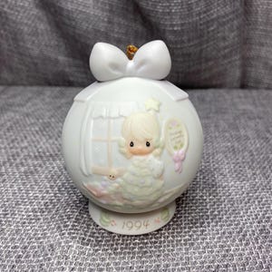 May include: A white porcelain Christmas ornament featuring a Precious Moments figurine of a girl holding a star. The ornament is decorated with a white bow and the year 1994. The text "Christmas Wishes" is printed on the ornament.