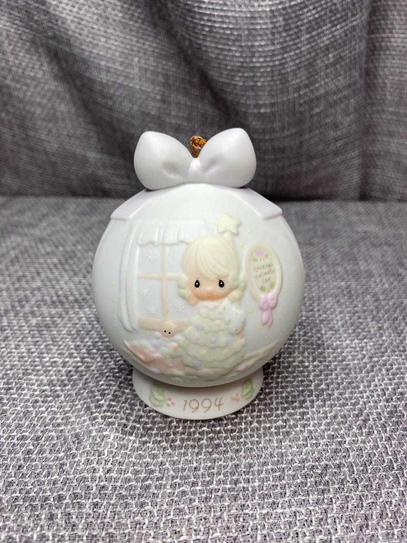 May include: A white porcelain Christmas ornament featuring a Precious Moments figurine of a girl holding a star. The ornament is decorated with a white bow and the year 1994. The text "Christmas Wishes" is printed on the ornament.