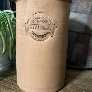Peut inclure: Une citerne à vin en grès de couleur beige avec un couvercle verni marron. L'avant de la citerne présente un emblème en relief avec les mots "Rowe Pottery Works" et "Wine Cistern". Le récipient cylindrique est conçu pour stocker du vin.