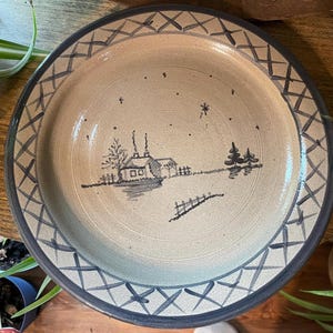 Puede incluir: Un plato redondo de cerámica con una escena invernal pintada a mano. El plato presenta una casa, árboles y una valla en una paleta de colores apagados. El borde tiene un patrón geométrico azul. Una pieza decorativa para el hogar.