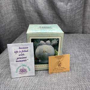May include: A blue porcelain Christmas tree ornament with a white bow and a small white bird perched on a branch. The ornament is part of the Precious Moments collection and features the text "You're As Pretty As A Christmas Tree 1994 Annual Edition".
