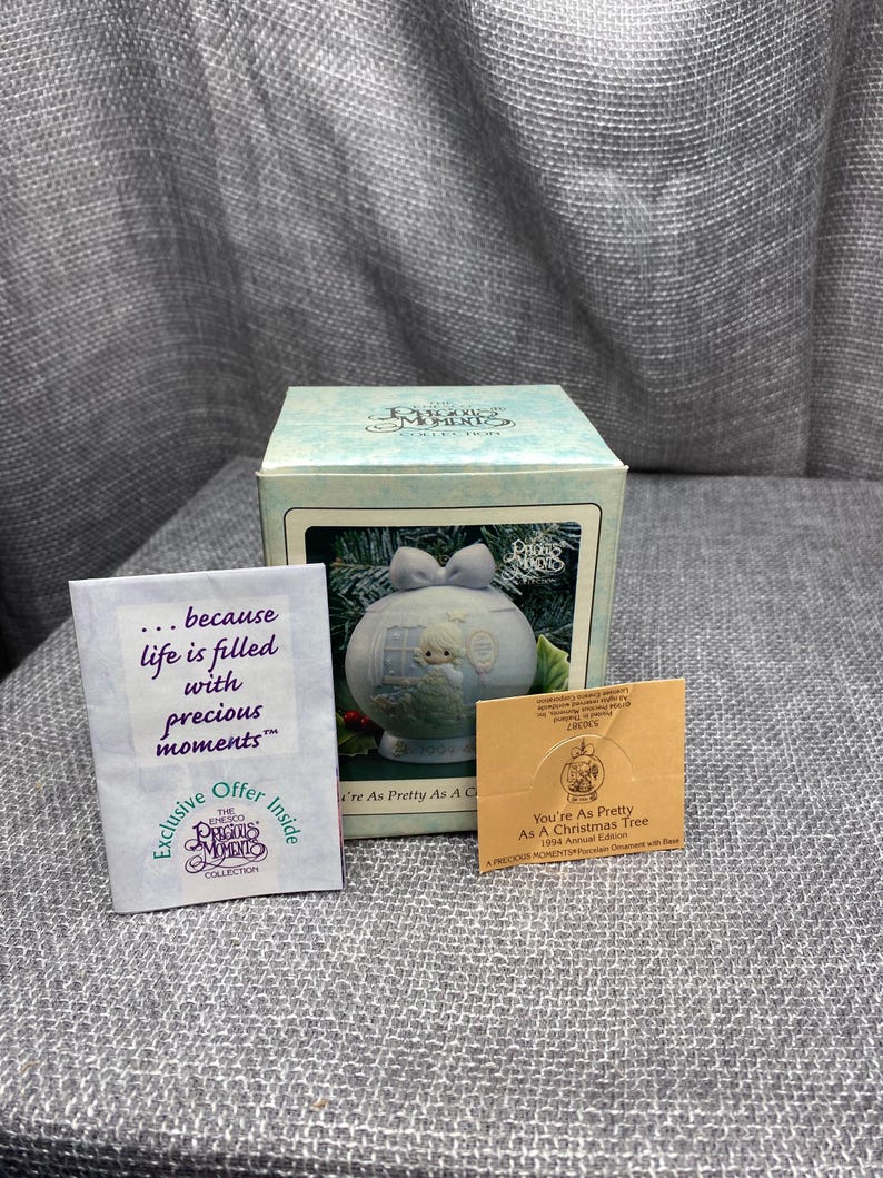 May include: A blue porcelain Christmas tree ornament with a white bow and a small white bird perched on a branch. The ornament is part of the Precious Moments collection and features the text "You're As Pretty As A Christmas Tree 1994 Annual Edition".