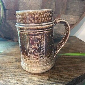 Pode incluir: Uma caneca de cerâmica marrom e branca com alça. A caneca tem um desenho de um prédio e o texto "Cambridge Pottery Festival 2004".