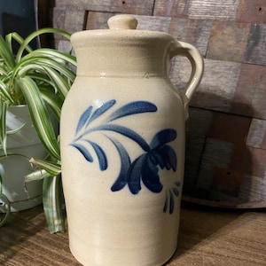 Op de afbeelding: Een crèmekleurige steengoedkan met een blauw bloemenmotief en een deksel. De kan heeft een handvat en is versierd met een blauw bloemenmotief.