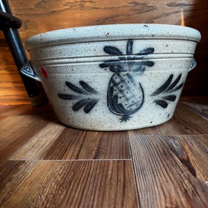 May include: A speckled beige stoneware bowl with a wide rim and two side handles. The exterior features a hand-painted black pineapple motif centered on the front, with a small red circular sticker visible on the left side near the handle.